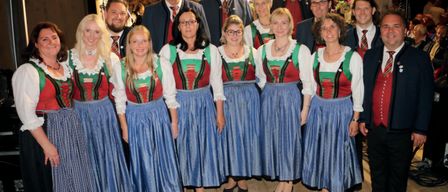 Eine Gruppe von Menschen in traditioneller deutscher Kleidung posiert für ein Foto vor einem festlichen Hintergrund, mit dem Datum 6. Dezember 2025 und dem Ort Im Gasthof.