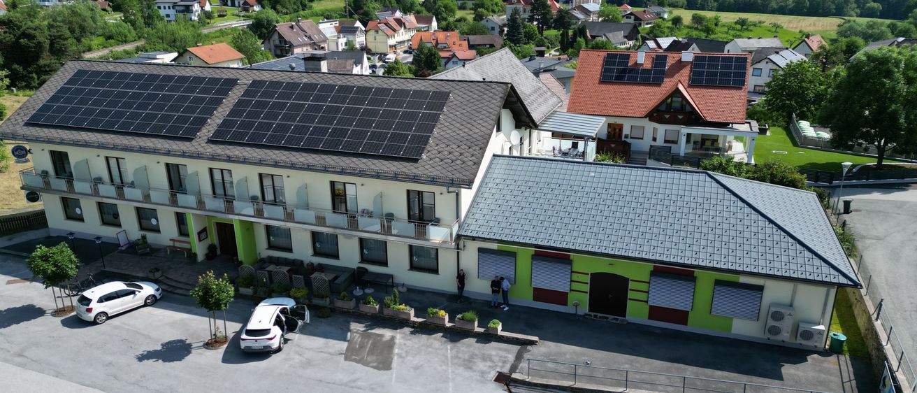 Luftaufnahme eines Dorfes mit Solarmodulen auf Häusern, grünen Bäumen und einem klaren blauen Himmel. Vor einem Gebäude mit Solarmodulen auf dem Dach sind einige Autos geparkt.