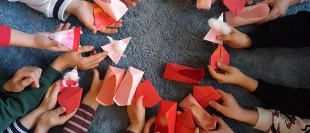 Eine Gruppe von Kindern sitzt im Kreis auf einem Teppich. Jedes Kind hält ein rotes Origami-Papierflugzeug in den Händen. Einige Flugzeuge haben Baumwolle darin. Die Hände der Kinder sind verschwommen.