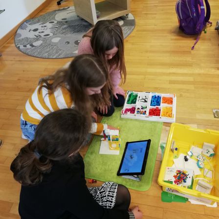 Drei Mädchen sitzen auf dem Boden und arbeiten an einem Projekt mit Legos und einem Tablet.
