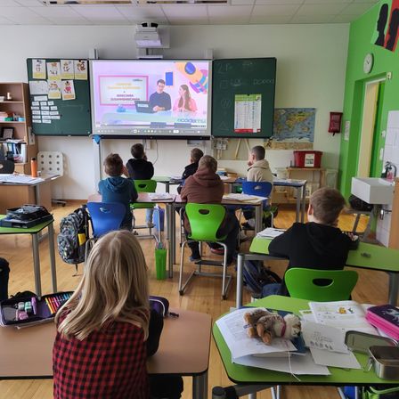 Ein Klassenzimmer mit Schülern in grünen Stühlen um Tische herum, die eine Präsentation auf einer großen Leinwand beobachten. Verschiedene Poster und ein Kalender hängen an der Tafel.