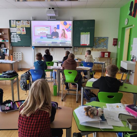 Ein Klassenzimmer mit Schülern in grünen Stühlen um Tische herum, die eine Präsentation auf einer großen Leinwand beobachten. Verschiedene Poster und ein Kalender hängen an der Tafel.
