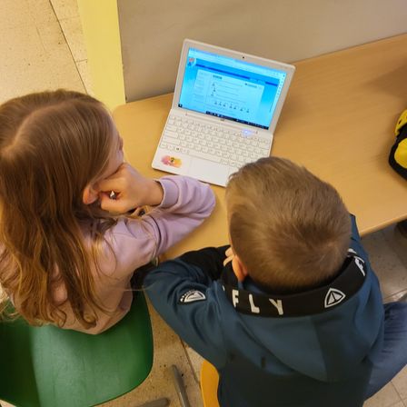 Zwei Kinder sitzen auf Stühlen, eines mit langen Haaren, die das Gesicht bedecken, das andere trägt eine blaue Kapuzenjacke mit schwarzem Kragen. Sie konzentrieren sich auf einen weißen Laptop mit blauem Bildschirm auf einem Holztisch.
