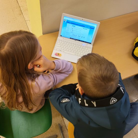 Zwei Kinder sitzen auf Stühlen, eines mit langen Haaren, die das Gesicht bedecken, das andere trägt eine blaue Kapuzenjacke mit schwarzem Kragen. Sie konzentrieren sich auf einen weißen Laptop mit blauem Bildschirm auf einem Holztisch.