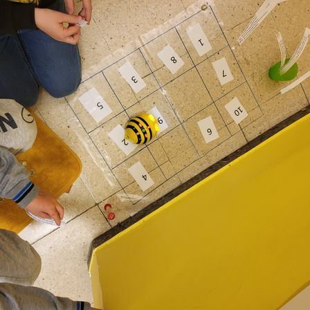 Zwei Kinder sitzen auf dem Boden und spielen ein Spiel mit einem bienenförmigen Spielzeug, einem durchsichtigen Spielbrett und einer gelben Box.