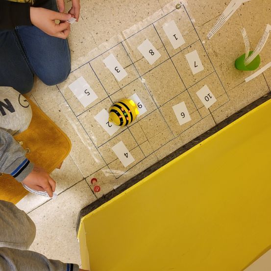 Zwei Kinder sitzen auf dem Boden und spielen ein Spiel mit einem bienenförmigen Spielzeug, einem durchsichtigen Spielbrett und einer gelben Box.