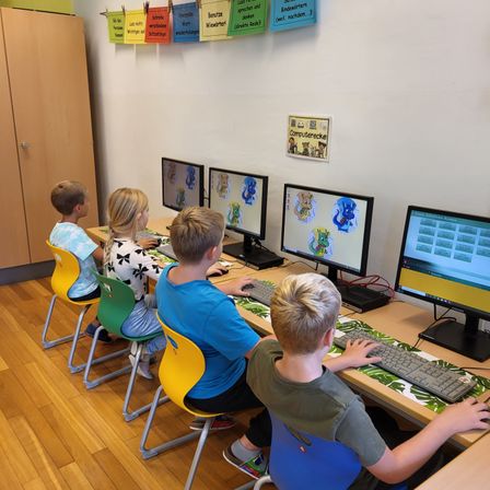 Vier Kinder sitzen an einem Computertisch, jedes benutzt einen Computer. Der Tisch hat mehrere Monitore, Tastaturen und Mäuse. Ein Schild an der Wand sagt 'Computerecke.'