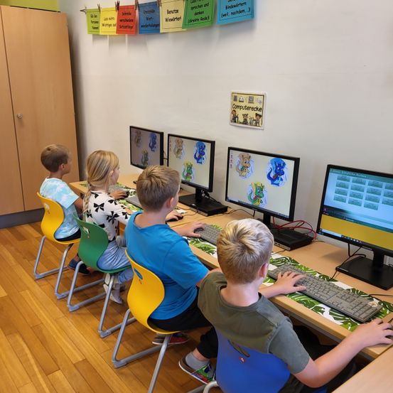 Vier Kinder sitzen an einem Computertisch, jedes benutzt einen Computer. Der Tisch hat mehrere Monitore, Tastaturen und Mäuse. Ein Schild an der Wand sagt 'Computerecke.'