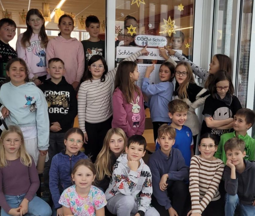 Eine Gruppe junger Schüler posiert für ein Foto im Klassenzimmer, einige halten ein Schild hoch, während andere für die Kamera lächeln.