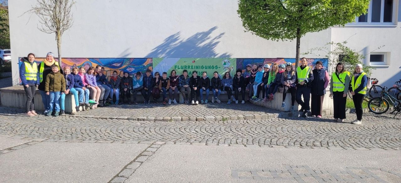 Eine Gruppe von Kindern sitzt zusammen auf einer Bank draußen mit einem Wandbild dahinter. Einige tragen Helme und einer hat ein grünes Schild.
