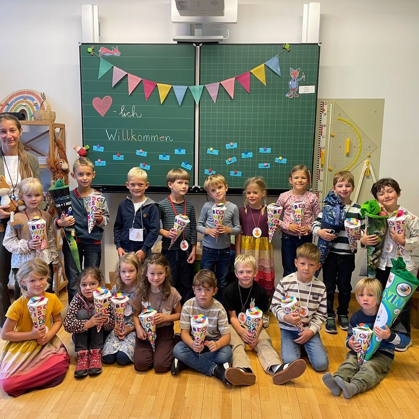 Ein Klassenzimmer voller Kinder, die dekorierte Kegel halten, mit einer Lehrerin vor einer Tafel. Der Raum ist mit bunten Wimpelketten und einem Banner geschmückt.