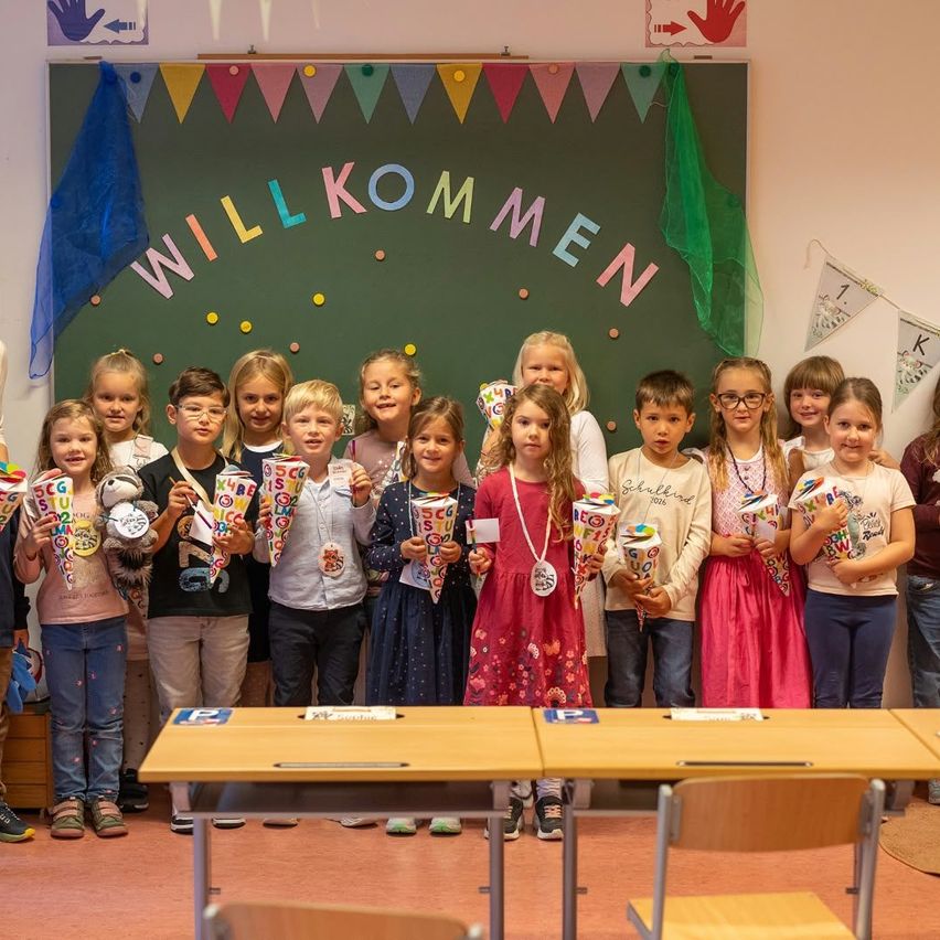 Eine Gruppe junger Kinder in einem Klassenzimmer, die Eistüten halten, steht vor einer Tafel mit bunten Dekorationen und dem Wort 'Willkommen' darauf.