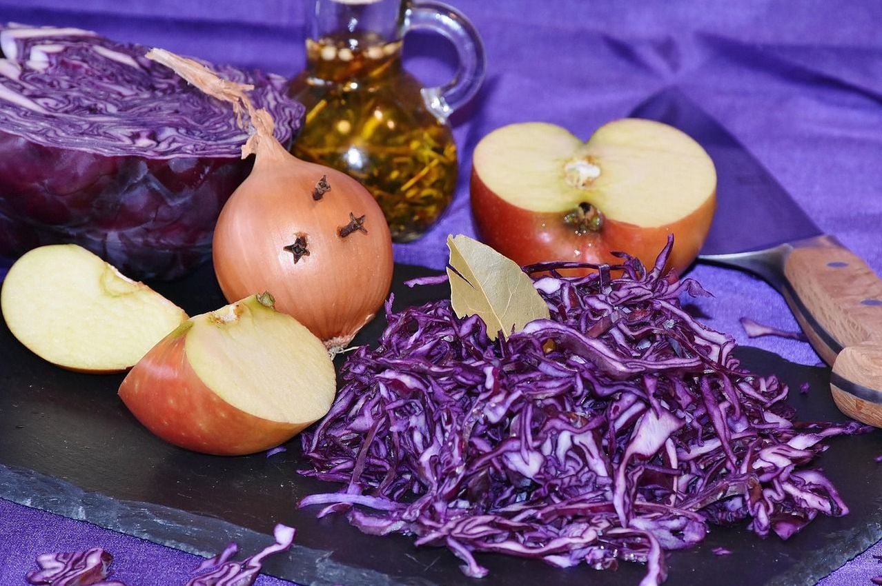 Ein Nahaufnahme eines Schneidebretts mit geschnittenem Rotkohl, einem halben Apfel, einer geschnittenen Zwiebel und einer Flasche Öl auf einem lila Tuch.