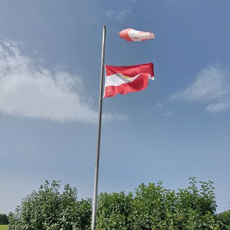 Eine rot-weiße Flagge weht im Wind auf einem Metallpfahl, mit einem rosa Ballo in der Nähe und Grünpflanzen darunter unter einem blauen Himmel mit Wolken.