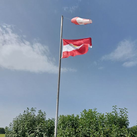 Eine rot-weiße Flagge weht im Wind auf einem Metallpfahl, mit einem rosa Ballo in der Nähe und Grünpflanzen darunter unter einem blauen Himmel mit Wolken.