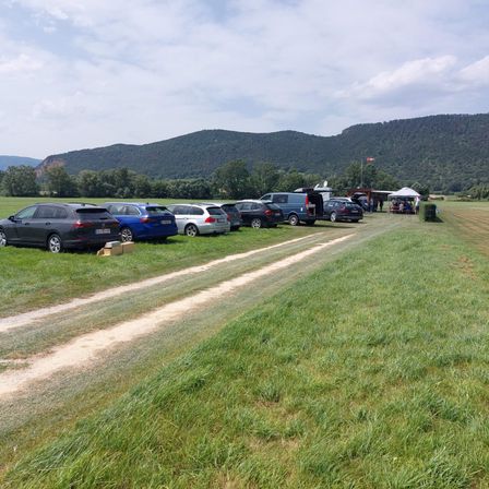 Ein grasbewachsenes Feld mit mehreren geparkten Autos, einem Feldweg und einem Zelt mit Leuten. Im Hintergrund befinden sich Berge und Bäume.