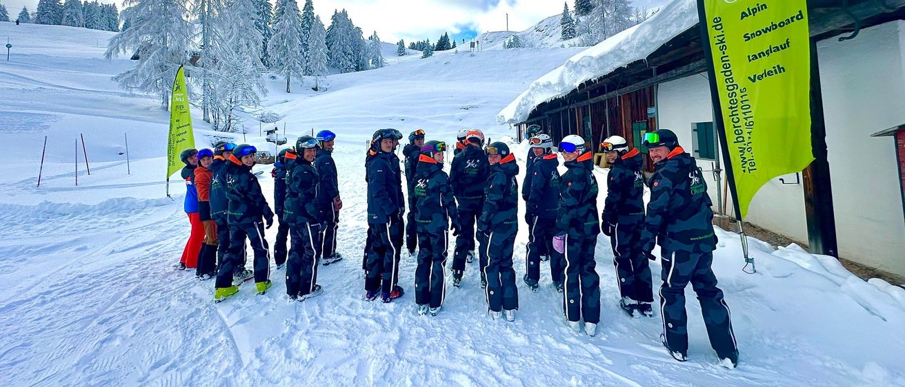 Eine Gruppe von Menschen in Winterkleidung steht auf einem verschneiten Feld, möglicherweise für ein Gruppenfoto. Sie sind von Tannenbäumen und einem verschneiten Berg umgeben. Banner befinden sich an der Seite.