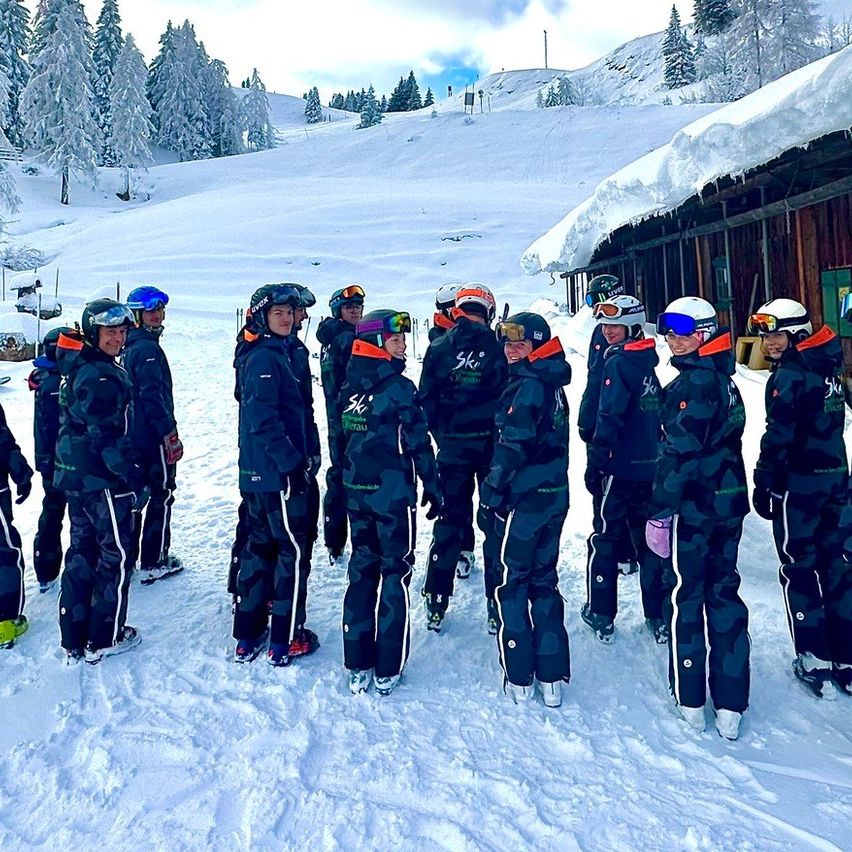 Eine Gruppe von Menschen in Winterkleidung steht auf einem verschneiten Feld, möglicherweise für ein Gruppenfoto. Sie sind von Tannenbäumen und einem verschneiten Berg umgeben. Banner befinden sich an der Seite.