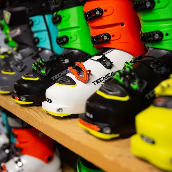 Ein Holzregal zeigt eine Reihe farbiger Skischuhe, darunter schwarz, grün, gelb, orange und türkis. Die Schuhe haben verschiedene Größen, und das Wort Tecnica ist auf dem weißen Schuh sichtbar.