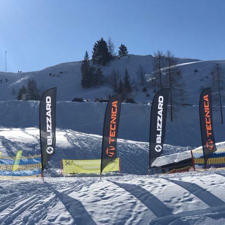 Mehrere Schneebanner stehen auf einer Skipiste mit Schneezäunen, Bäumen und einem Berg im Hintergrund. Die Banner tragen Blizzard, Tecnica und Skigebietsmarken.