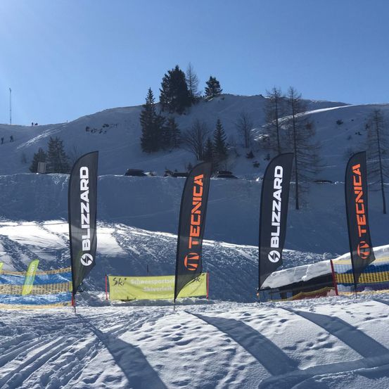 Mehrere Schneebanner stehen auf einer Skipiste mit Schneezäunen, Bäumen und einem Berg im Hintergrund. Die Banner tragen Blizzard, Tecnica und Skigebietsmarken.