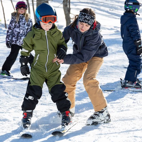 Ein junges Kind lernt Skifahren mit einer erwachsenen Helferin an einem verschneiten Tag. Das Kind trägt einen grünen Skianzug und eine Brille. Die Helferin, eine Frau, trägt eine blaue Jacke und braune Hose. Zwei weitere Kinder sind im Hintergrund beim Skifahren.