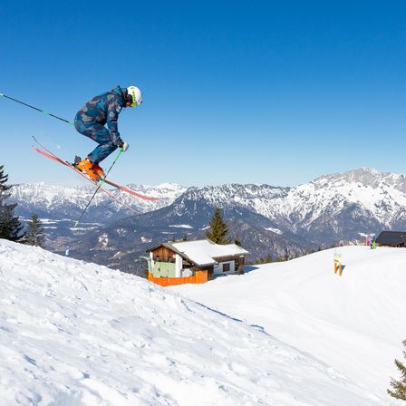 Ein Skifahrer in einem blauen und schwarzen Outfit ist über einem verschneiten Berg in der Luft. Verschneite Gipfel sind im Hintergrund zu sehen. Ein kleines Chalet ist auf der rechten Seite sichtbar.