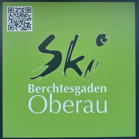Ein grünes Poster mit 'Ski' in stilisierter Schrift und 'Berchtesgaden Oberau' darunter. Ein QR-Code befindet sich in der oberen linken Ecke.