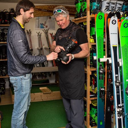 Zwei Männer in einem Skigeschäft, einer hält einen Skischuh, während der andere ihn untersucht. Der Laden ist mit Skiern und Snowboards gefüllt.