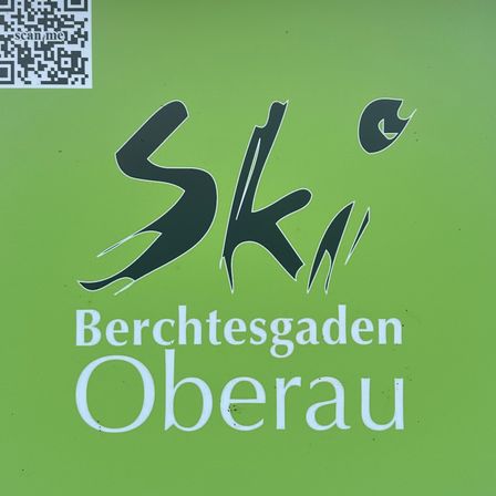Ein grünes Plakat mit einem QR-Code und stilisiertem Text, der 'Ski Berchtesgaden Oberau' lautet. Der QR-Code befindet sich auf der linken Seite.