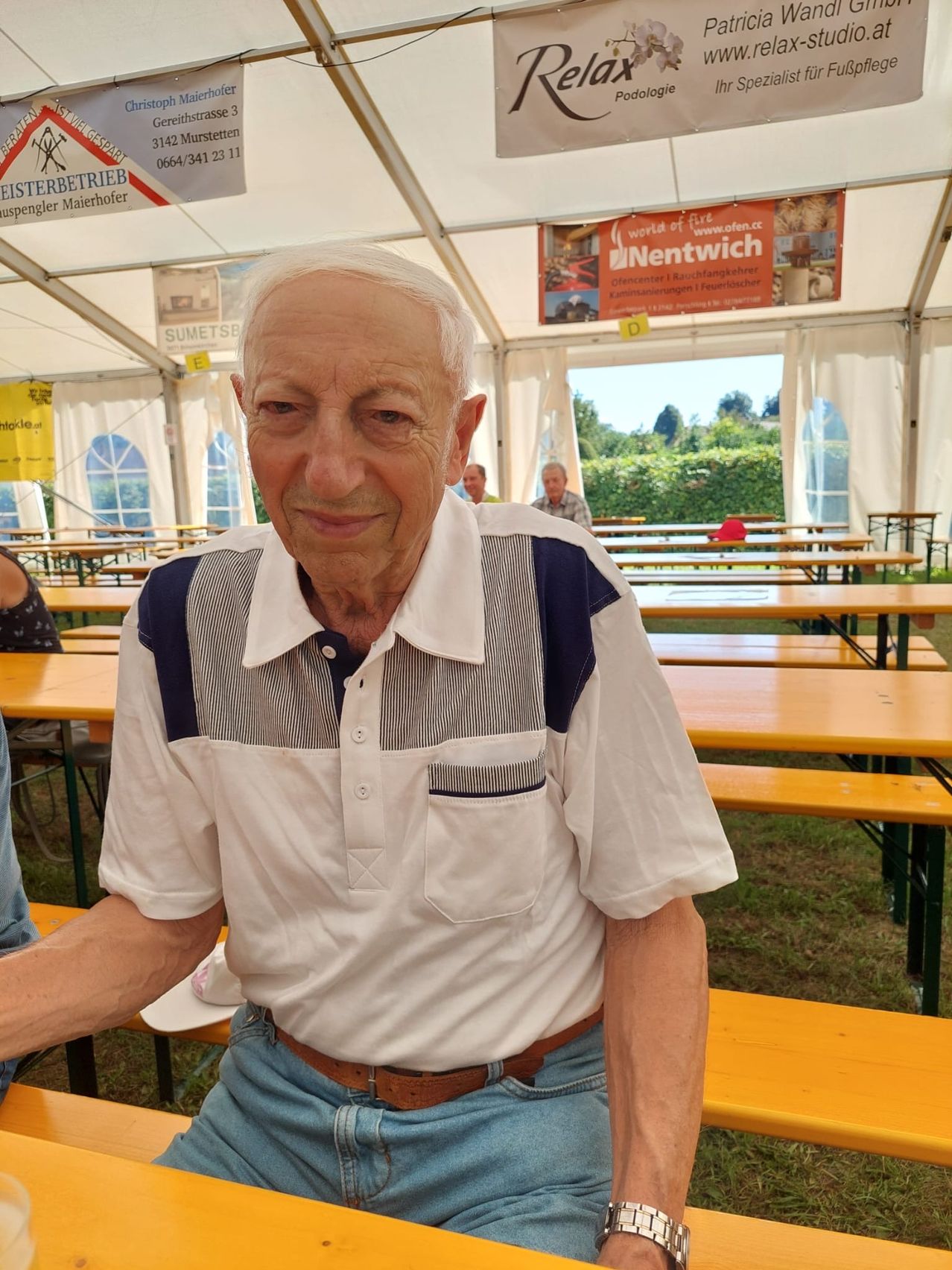 Ein älterer Mann in einem weißen Hemd mit blauem Streifen sitzt an einem Tisch unter einem Zelt und lächelt in die Kamera. Hinter ihm sind weitere Tische und Bänke zu sehen, an denen Menschen sitzen und stehen.