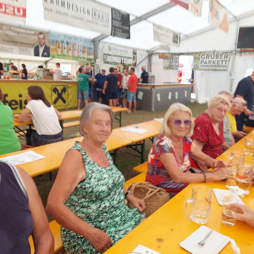Eine Gruppe älterer Frauen sitzt an einem gelben Tisch in einem Zelt. Sie trinken aus Gläsern. Ein Mann in Anzug ist auf einem Banner abgebildet.