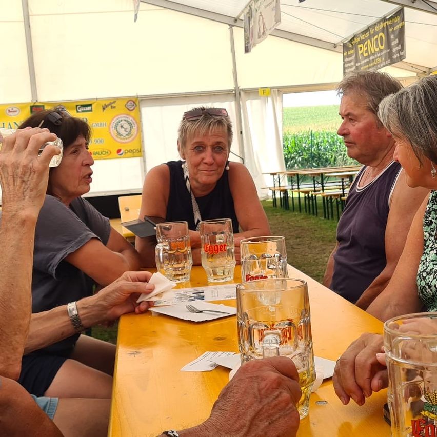 Eine Gruppe von Menschen sitzt an einem gelben Tisch mit Bierkrügen. Eine Frau trinkt aus einem Glas, während andere sich unterhalten. Hinter ihnen ist ein weißes Zelt und ein gelber Banner zu sehen.