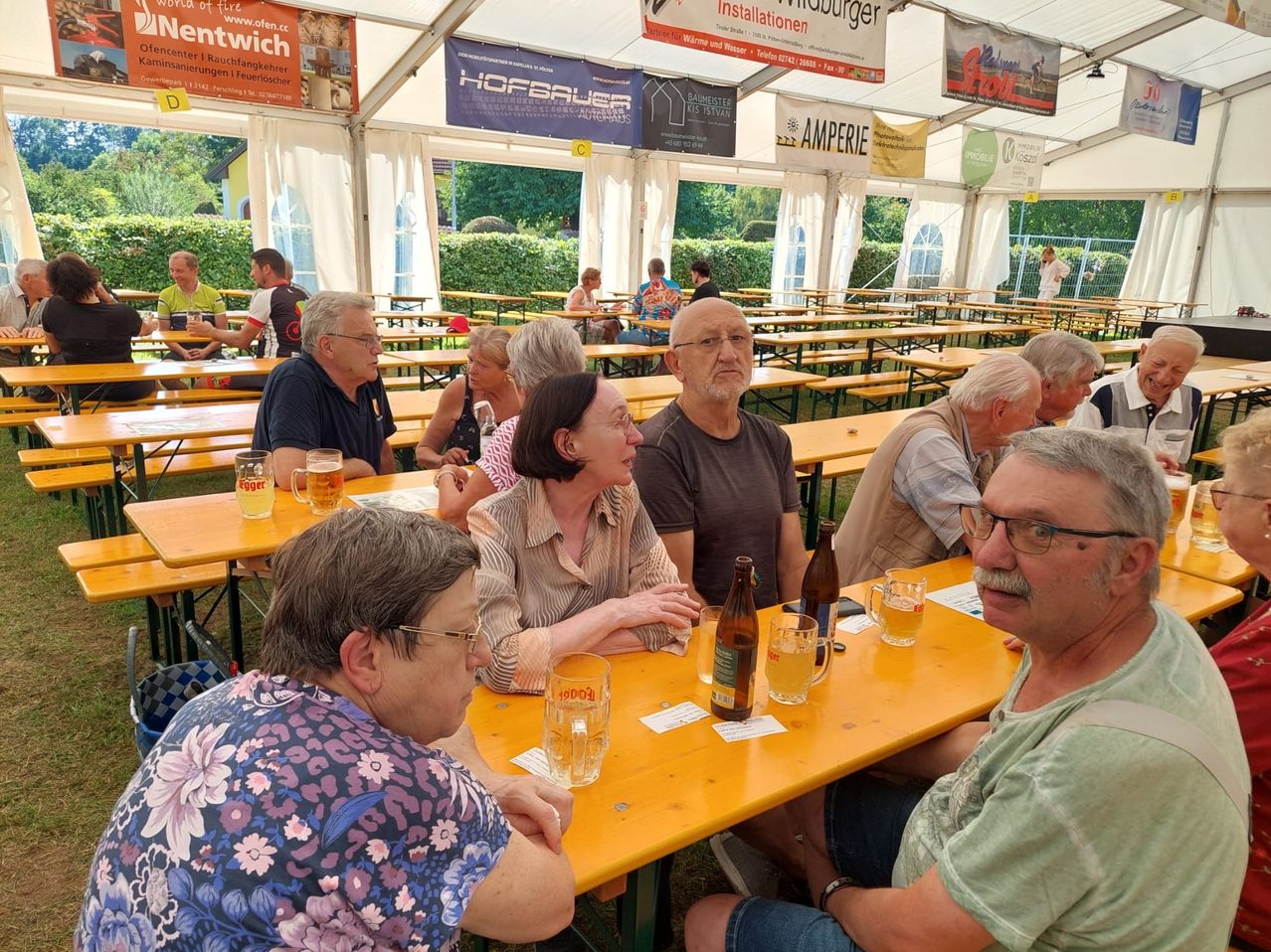 Eine Gruppe von Menschen sitzt an Tischen in einem Zelt und genießt Getränke und Essen. Banner hängen über ihnen, und einige tragen Brillen und Uhren. Bäume und ein Gebäude sind im Hintergrund zu sehen.