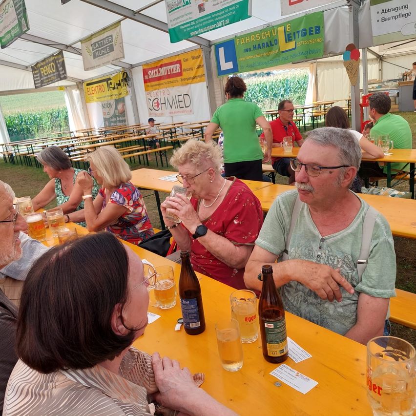 Mehrere ältere Menschen sitzen um einen Holztisch herum und trinken Bier aus Gläsern. Einige tragen Brillen und Uhren. Eine Frau in einem roten Shirt trinkt aus einem Glas. Ein Schild hängt über den Tischen.