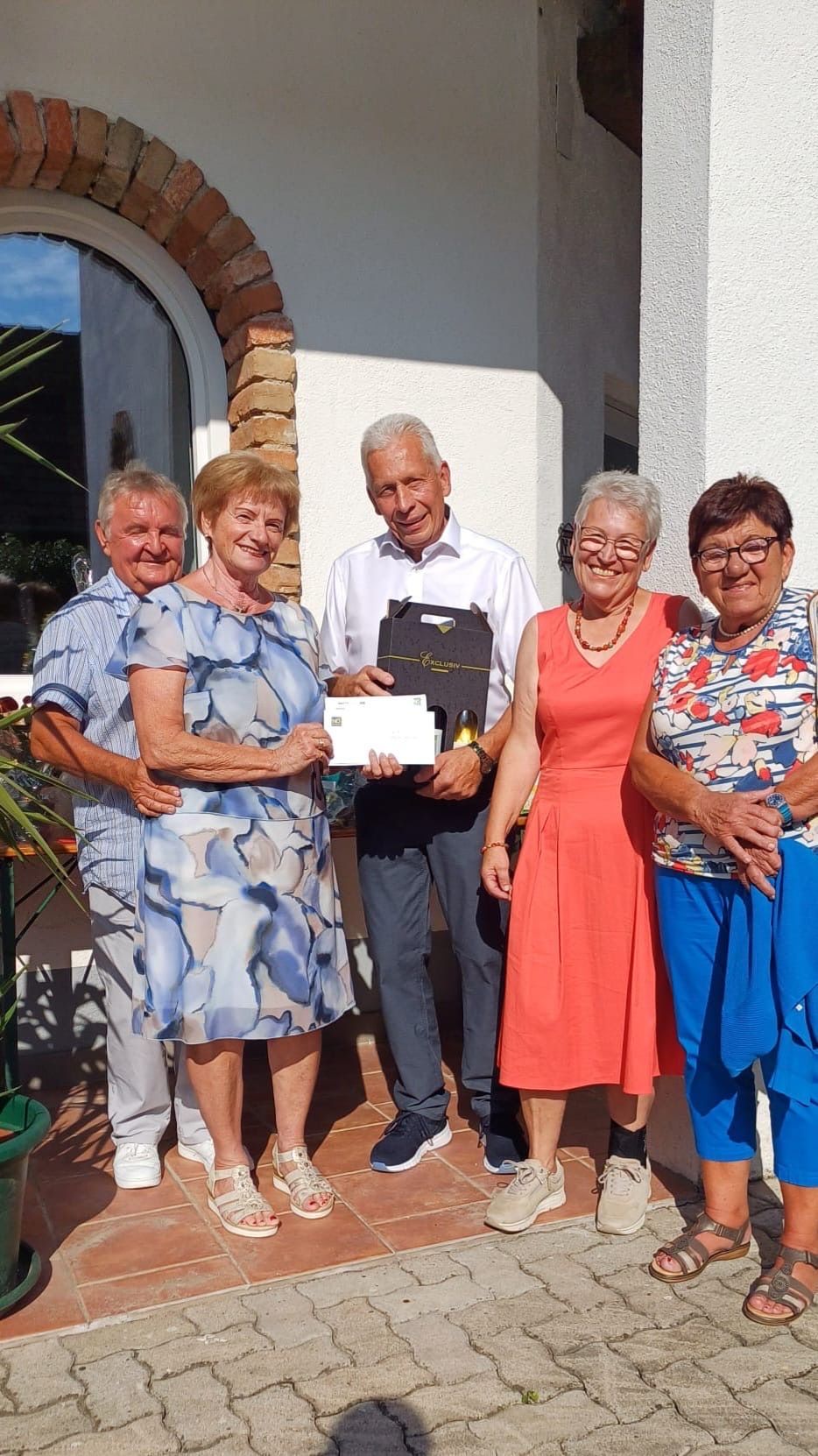 Fünf Menschen stehen lächelnd zusammen auf einer Veranda. Sie halten ein Zertifikat und eine Geschenkbox. Hinter ihnen stehen eine Pflanze und ein Glasfenster.