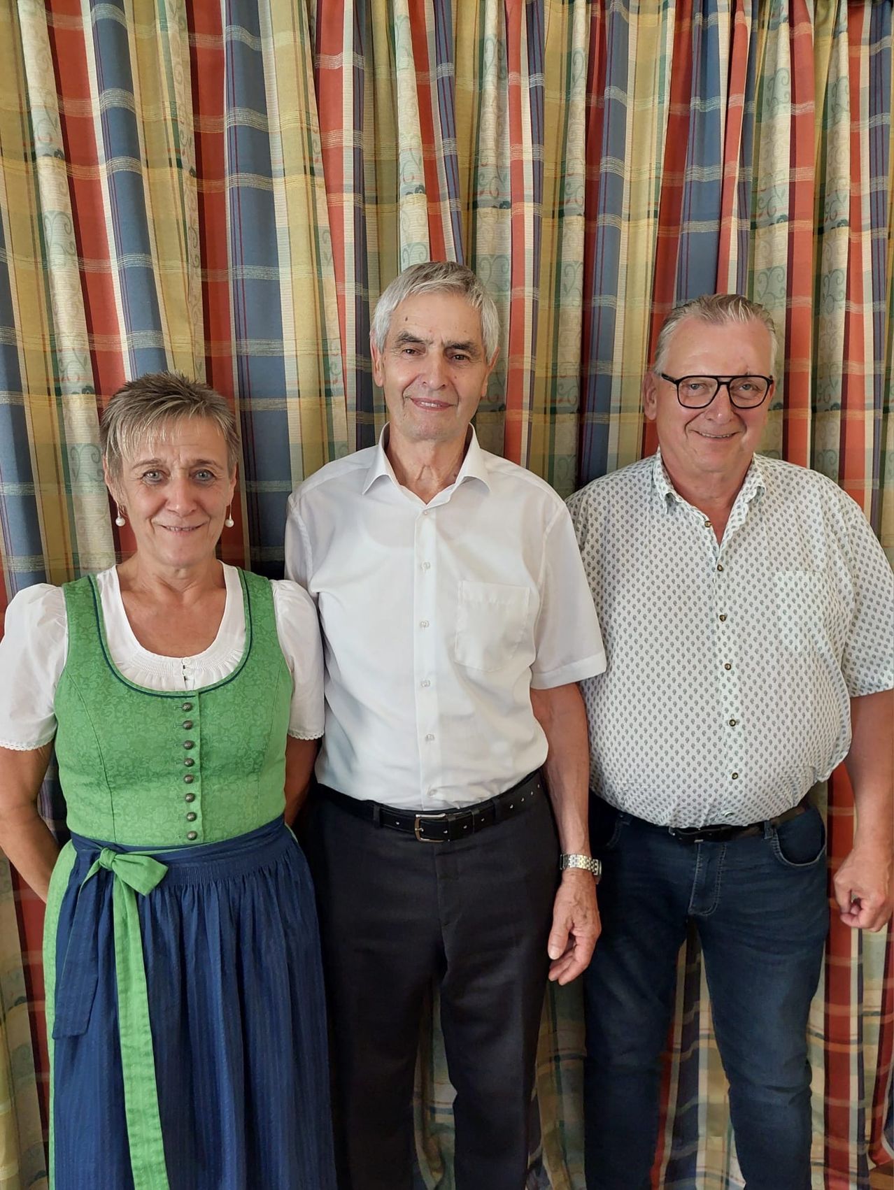 Drei Menschen stehen lächelnd nebeneinander, Frau in grünem Kleid, zwei Männer in weißen Hemden, einer mit Brille. Sie stehen vor einem farbenfrohen Vorhang.