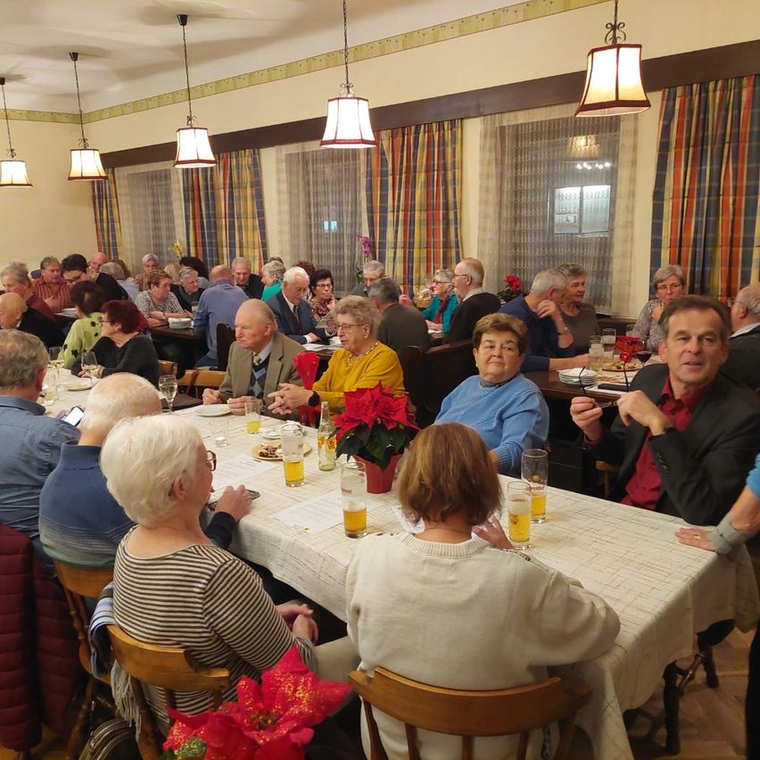 Eine Gruppe älterer Menschen diniert an einem langen Tisch mit Getränken und Blumen auf dem Tisch. Sie unterhalten sich.
