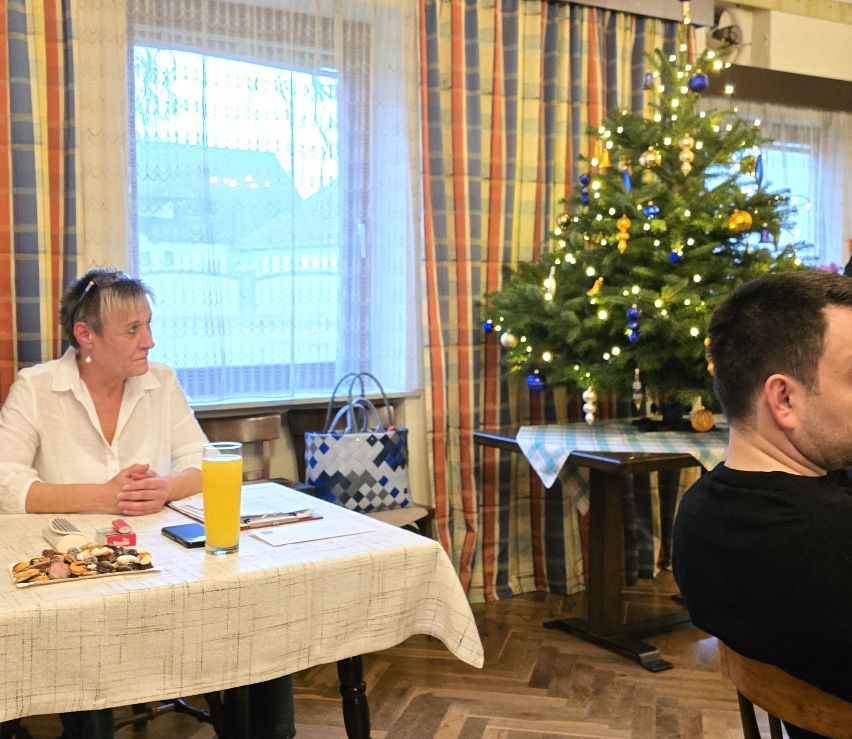 Eine Frau in einem weißen Hemd sitzt an einem Tisch mit einem Glas Orangensaft. Ein Mann sitzt ihr gegenüber. Dahinter steht ein mit Lichtern geschmückter Weihnachtsbaum.