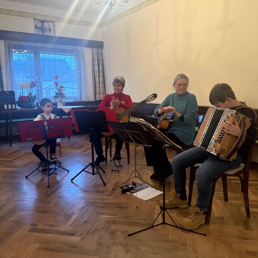 Drei Erwachsene und zwei Kinder sitzen auf Stühlen und spielen Instrumente, wahrscheinlich in einem Musikraum. Die Erwachsenen spielen Gitarren und ein Akkordeon. Die Kinder, die vorne sitzen, scheinen Klavier zu spielen. Ein Notenständer hält Notenblätter.