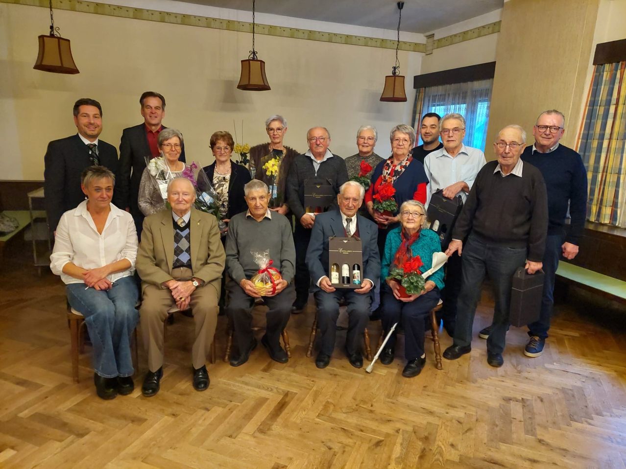 Eine Gruppe von Menschen, meist Senioren, posiert für ein Foto in einem Raum mit Holzböden. Einige halten Blumen, Geschenke und eine Schachtel. Sie alle lächeln und scheinen ein Ereignis zu feiern.