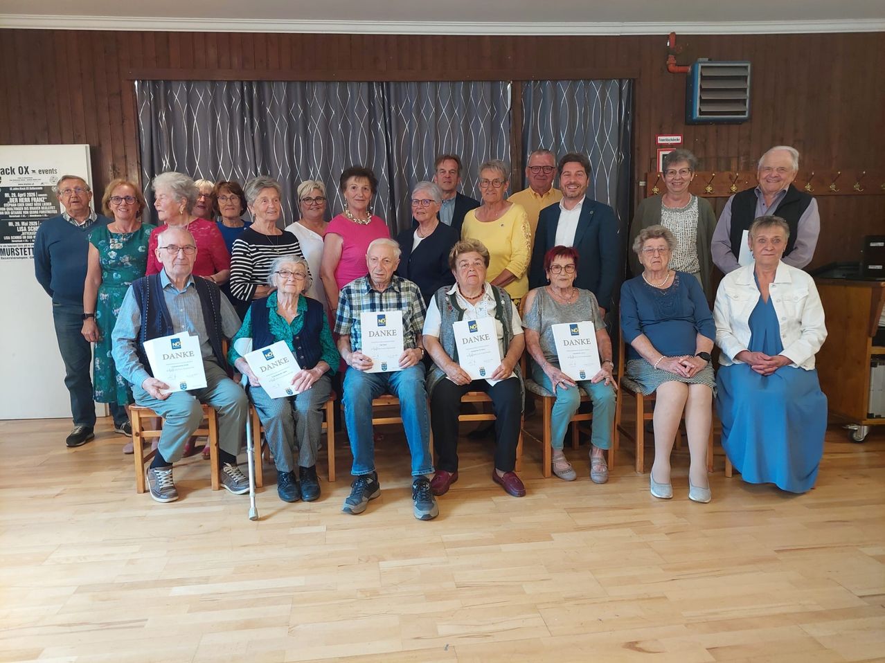 Eine Gruppe älterer Menschen, die Urkunden halten, posiert für ein Foto in einem Raum mit Holzwänden und -böden. Sie sitzen auf Stühlen und einige haben Brillen.