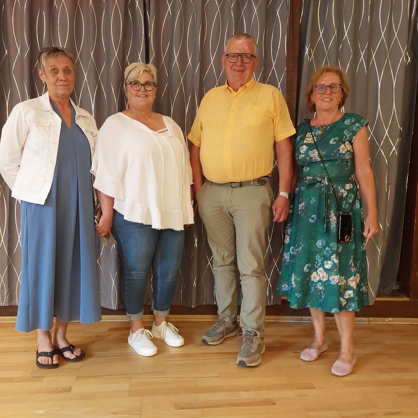 Vier Menschen stehen gemeinsam auf einem Holzboden und lächeln für ein Foto vor einem Vorhang. Der Mann trägt ein gelbes Hemd und die Frauen tragen eine Jacke, ein Blumenkleid und blaue Kleider.