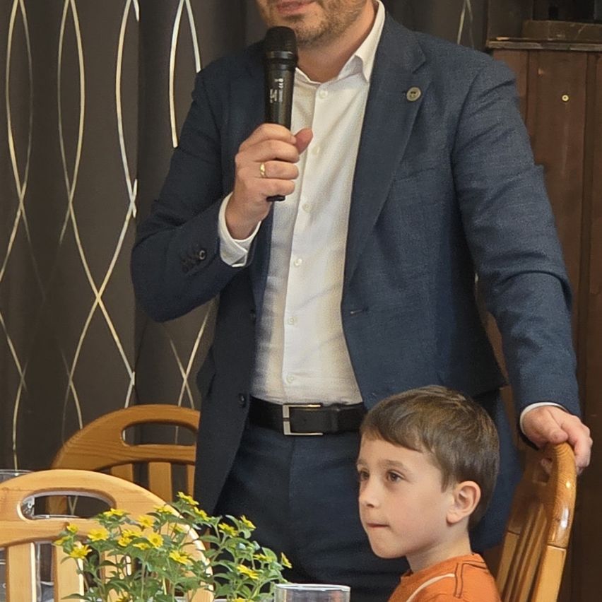 Ein Mann steht hinter einem Mikrofon und spricht in dieses. Ein Junge sitzt davor und hört aufmerksam zu. Sie befinden sich in einem Innenraum mit dekorativem Tapetenmuster und Holzstühlen.