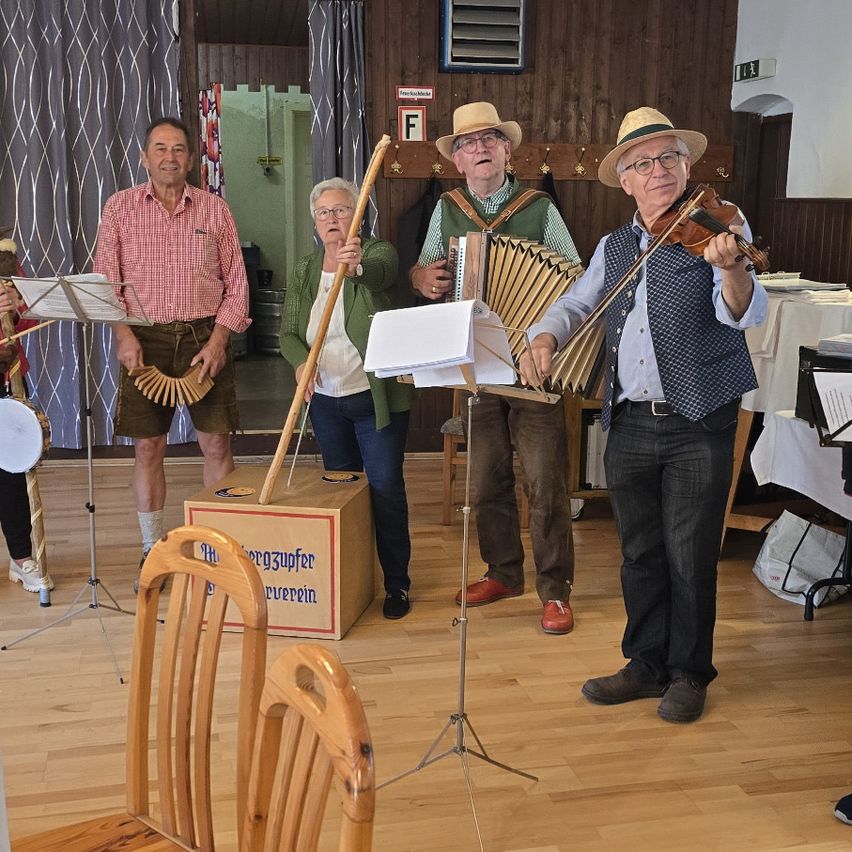 Fünf Menschen spielen Musik in einem Raum, zwei Männer mit Violinen, eine Frau mit einer Flöte und zwei Männer mit Akkordeons. Der Raum hat Holzböden und -wände.
