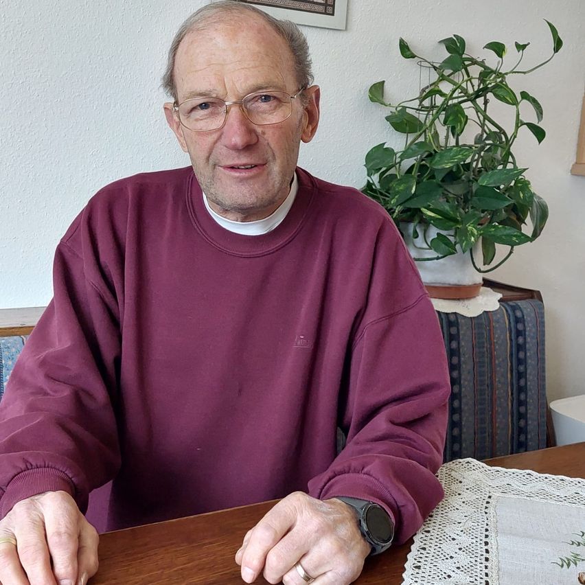 Ein älterer Mann mit einer Brille, der einen Weinroten Pullover trägt, sitzt an einem hölzernen Tisch mit einer Spitzentischdecke, mit einem Topfpflanzen hinter ihm.