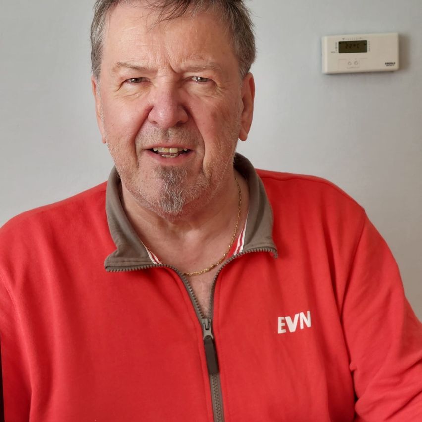 Ein Mann in einem roten Jacke mit den Buchstaben EVN darauf, lächelt und sieht in die Kamera. Ein Thermostat ist an der Wand hinter ihm angebracht.
