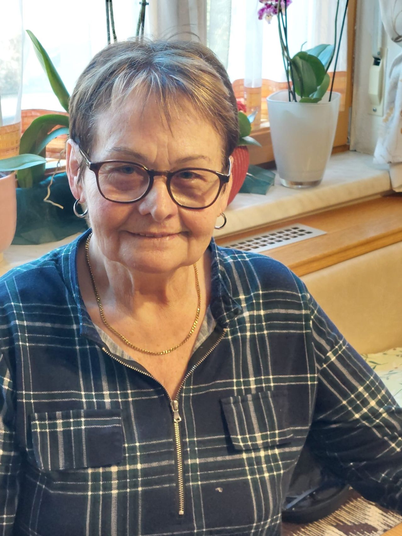 Eine ältere Frau mit kurzen Haaren, Brille und blauem kariertem Shirt mit Reißverschluss sitzt an einem Tisch mit Topfpflanzen im Hintergrund.
