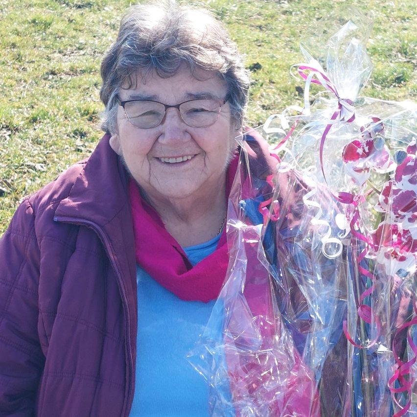 Eine lächelnde Frau mit Brille hält einen Blumenstrauß, der in Plastik eingewickelt ist. Sie trägt eine lila Jacke und einen rosa Schal. Der Ort ist eine Rasenfläche.