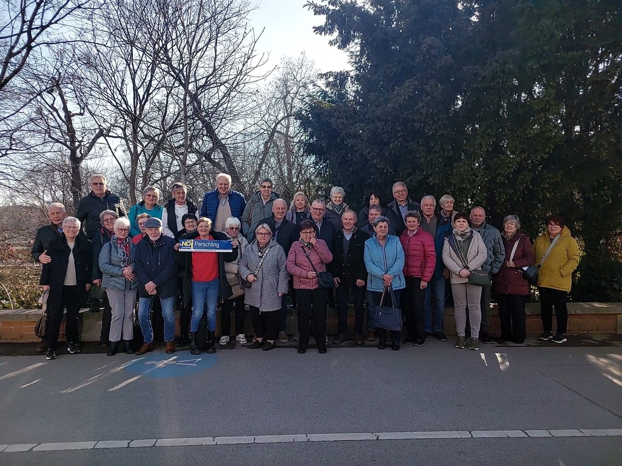 Eine Gruppe Erwachsener versammelt sich im Freien, posiert für ein Foto vor Bäumen und einer Straße. Eine Person hält ein blaues Schild mit der Aufschrift Perschiling.
